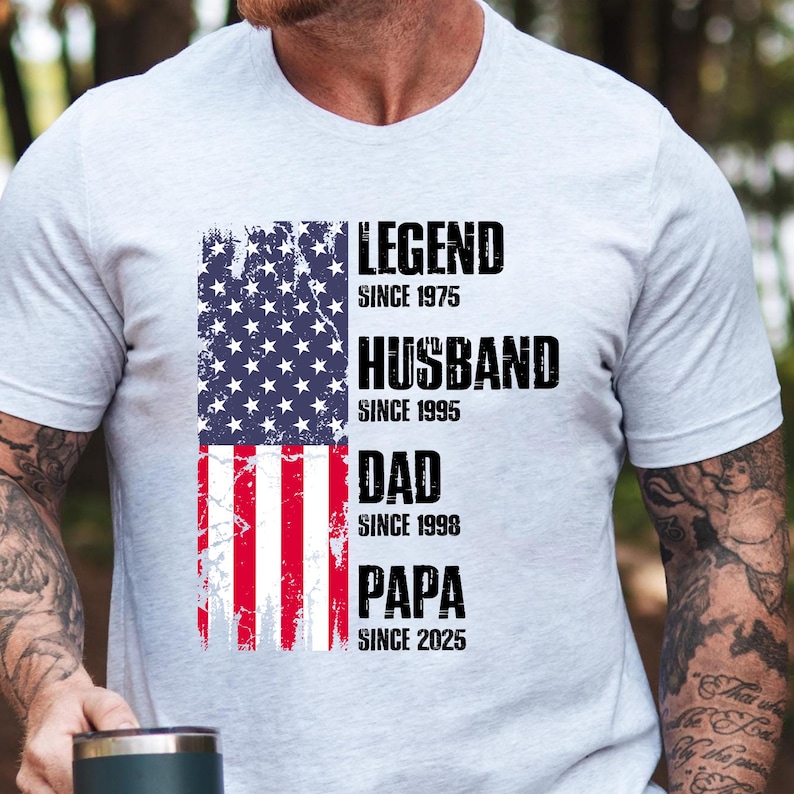 K&ouml;nnte beinhalten: Ein hellgraues T-Shirt mit einem verblassten amerikanischen Flaggen-Design. Das Shirt zeigt die Worte "LEGEND SINCE 1975", "HUSBAND SINCE 1995", "DAD SINCE 1998" und "PAPA SINCE 2025" in fetter schwarzer Schrift.