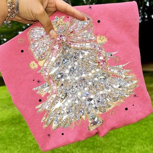 以下が含まれることがあります： ピンク色の装飾用クッション。キラキラ光るクリスマスツリーのデザイン。ツリーは大きなリボンなど、シルバーとゴールドのアクセントで飾られています。黒い星型の装飾がデザインの周りに散りばめられています。