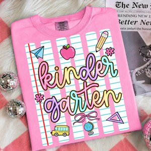 Peut inclure: T-shirt rose Comfort Colors avec un motif "Kindergarten" color&eacute;. Le design comprend un avion en papier, une pomme, un crayon, une fleur, un n&oelig;ud, un bus scolaire, un globe et un triangle. Le t-shirt a un col rond et des manches courtes.