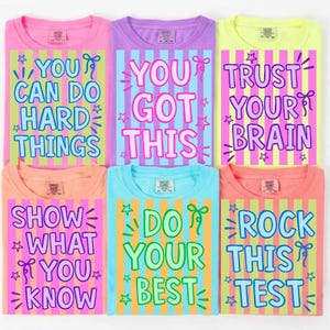 May include: A collection of six colorful t-shirts with positive affirmations. Each shirt features a different message in a playful font, set against a striped background. The shirts are in shades of pink, purple, yellow, peach, blue, and coral.