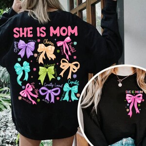 K&ouml;nnte beinhalten: Schwarzes Sweatshirt mit dem Text "SHE IS MOM" in rosa Buchstaben und einem Design aus bunten Schleifen mit W&ouml;rtern wie "Strong", "Chosen", "Beautiful", "Victorious", "Greatly Loved" und "Capable".