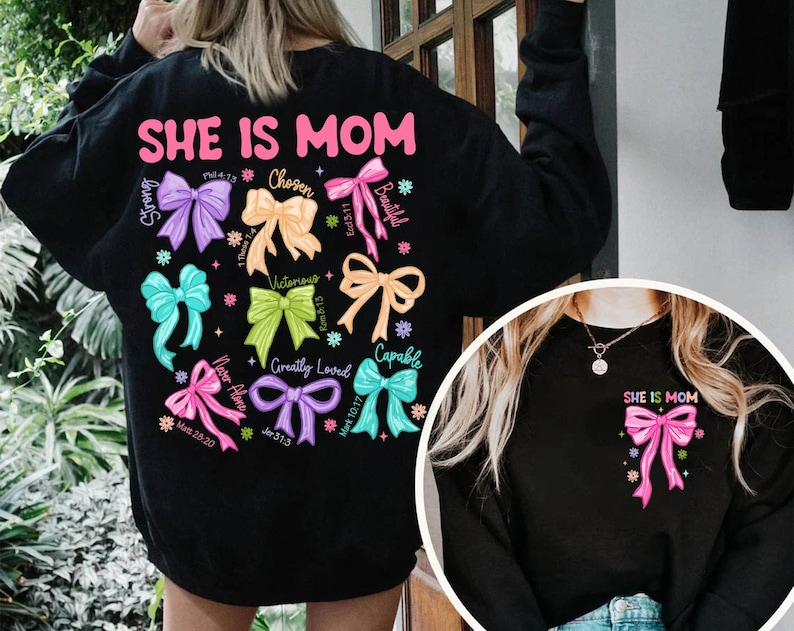K&ouml;nnte beinhalten: Schwarzes Sweatshirt mit dem Text "SHE IS MOM" in rosa Buchstaben und einem Design aus bunten Schleifen mit W&ouml;rtern wie "Strong", "Chosen", "Beautiful", "Victorious", "Greatly Loved" und "Capable".