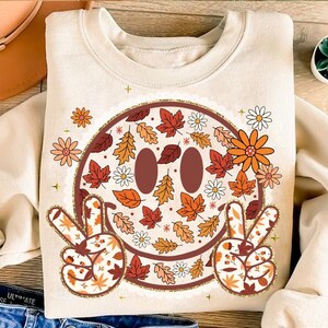 May include: Cream-colored sweatshirt featuring a smiley face design filled with autumn leaves and flowers in shades of orange, red, and brown. The smiley face has peace signs with leaf patterns. The sweatshirt has a round neckline.