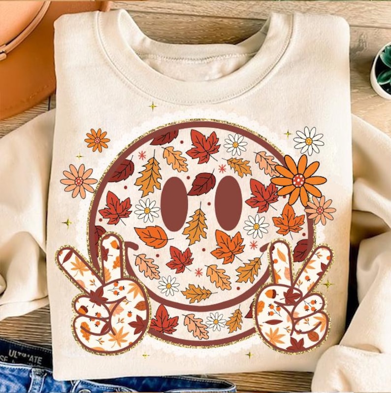May include: Cream-colored sweatshirt featuring a smiley face design filled with autumn leaves and flowers in shades of orange, red, and brown. The smiley face has peace signs with leaf patterns. The sweatshirt has a round neckline.