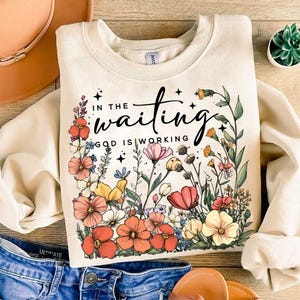 May include: A cream-colored sweatshirt with a floral design and the text "In the waiting, God is working".