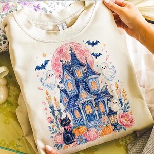 Puede incluir: Una sudadera de cuello redondo color crema con un estampado gr&aacute;fico de una casa encantada azul oscuro, una luna rosa grande, murci&eacute;lagos, fantasmas blancos, velas encendidas, calabazas naranjas, flores y un gato negro con un lazo rosa.