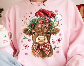 May include: A pink sweatshirt with a brown and white cow wearing a red and white Santa hat and a red and white plaid bow tie. The cow is surrounded by snowflakes, holly, and candy canes.