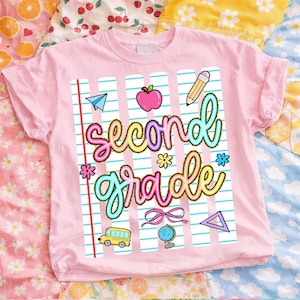 Peut inclure: T-shirt rose clair avec l'inscription "second grade" dans une police color&eacute;e et fantaisiste. Le motif comprend des graphiques sur le th&egrave;me de l'&eacute;cole, comme une pomme, un crayon, un avion en papier et un bus scolaire. Le t-shirt est en mati&egrave;re souple.