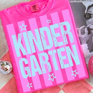 May include: A bright pink t-shirt with vertical stripes and the word "KINDERGARTEN" in light blue, along with star graphics. The shirt is made of a soft material and is likely for children.