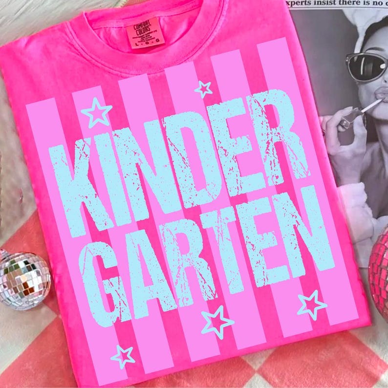 May include: A bright pink t-shirt with vertical stripes and the word "KINDERGARTEN" in light blue, along with star graphics. The shirt is made of a soft material and is likely for children.