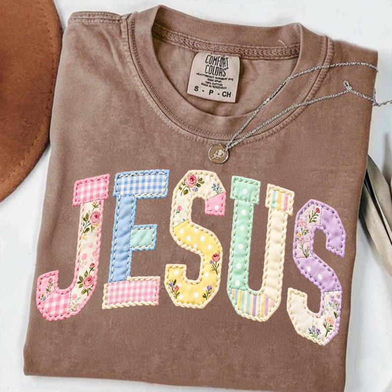 Pode incluir: Uma camiseta Comfort Colors marrom com a palavra "JESUS" aplicada na frente. As letras s&atilde;o feitas de v&aacute;rios tecidos coloridos em tons pastel, incluindo estampas florais e xadrez. Um colar de prata com um pingente est&aacute; sobre a camiseta.