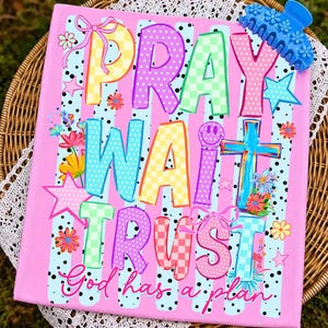 May include: Pink canvas art with the words "PRAY WAIT TRUST" in colorful, patterned letters. The artwork includes a cross, stars, flowers, and the phrase "God has a plan." A blue hair clip with flower details rests on the top right corner.