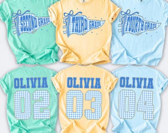 May include: Three t-shirts in mint, yellow, and light blue. The front features a blue gingham pennant with text reading SECOND GRADE, THIRD GRADE, and FOURTH GRADE. The back displays the name OLIVIA with the numbers 02, 03, and 04 in blue.
