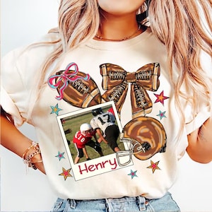 May include: White t-shirt with a brown football, a brown bow, a football helmet, and a photo of a football player. The photo has the name "Henry" written below it. The shirt has a colorful star and bow design.