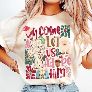 May include: A white t-shirt with a Christmas-themed design featuring a cross, a lamb, a star, and the text "Come Let Us Adore Him".