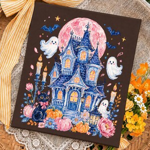 Puede incluir: Un lienzo rectangular con fondo marr&oacute;n oscuro que presenta una ilustraci&oacute;n detallada de una casa encantada azul, una luna rosa grande, tres fantasmas blancos, un gato negro con un lazo rosa, calabazas naranjas y velas encendidas.