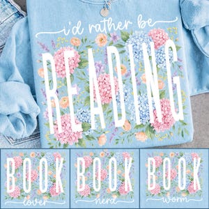 May include: Pale blue sweatshirt featuring the words "I'd rather be READING" in white, framed by a floral arrangement of pink, blue, and peach flowers. Beneath are three squares with the words "BOOK lover", "BOOK nerd", and "BOOK worm".