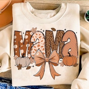 Puede incluir: Una sudadera beige con la palabra "mama" en letras marrones, naranjas y blancas con un diseño estampado. Las letras están decoradas con hojas de otoño, un lazo y dos calabazas.