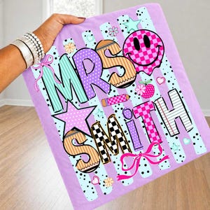 May include: A lavender blanket with the words "MRS SMITH" in colorful, playful lettering. The design includes a checkered smiley face, stars, hearts, and a bow. The background has light blue stripes and black polka dots.