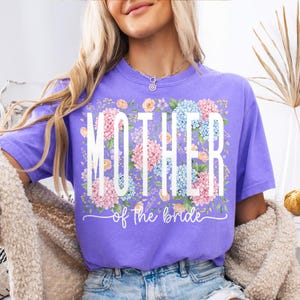 May include: A lilac-colored t-shirt with the words "MOTHER" and "of the bride" in white script. The word "MOTHER" is superimposed over a floral arrangement of pink and blue hydrangeas and other flowers.