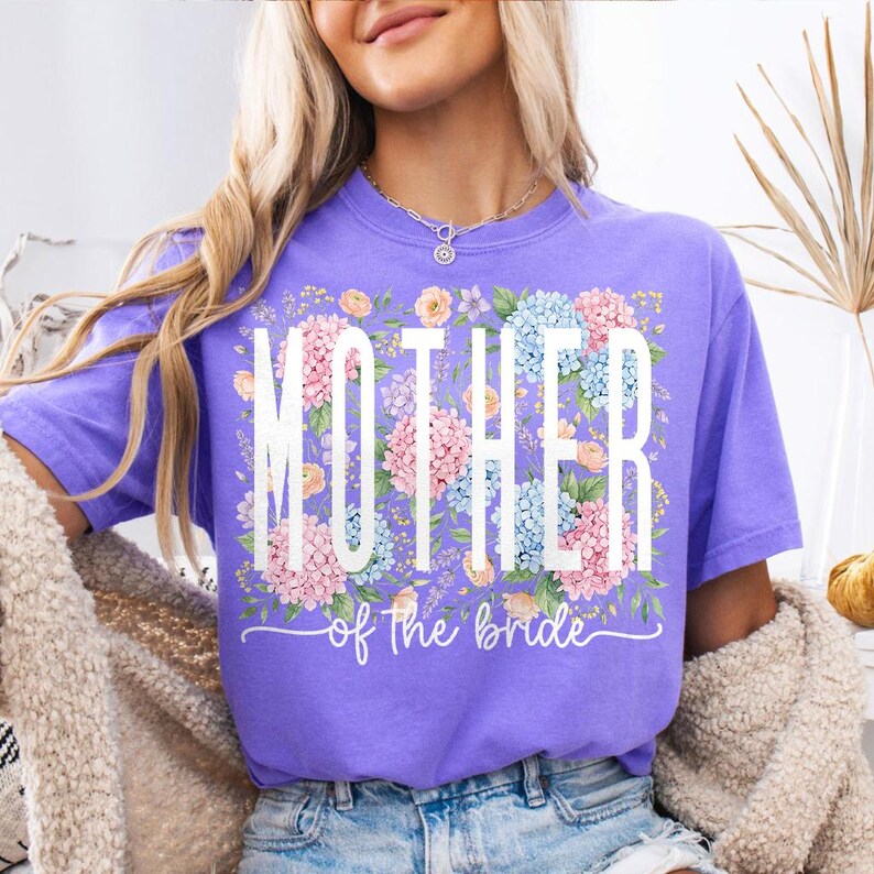 May include: A lilac-colored t-shirt with the words "MOTHER" and "of the bride" in white script. The word "MOTHER" is superimposed over a floral arrangement of pink and blue hydrangeas and other flowers.