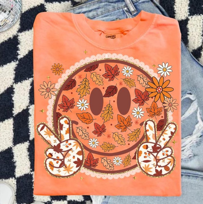 May include: Orange crew neck t-shirt featuring a smiley face design with fall leaves, daisies, and peace signs. The smiley face has brown eyes and is surrounded by a gold glitter trim. The shirt is folded on a denim surface.
