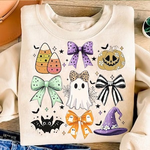 May include: A cream-colored sweatshirt with a Halloween-themed design. The design features a variety of cute Halloween icons, including candy corn, bows, a ghost, a witch's hat, a bat, and a pumpkin.