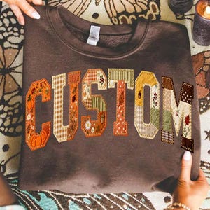 May include: A dark brown crewneck sweatshirt featuring the word CUSTOM in large, block-style letters. Each letter is filled with a unique patchwork pattern of autumn-themed fabrics, including pumpkins, acorns, leaves, and plaid in shades of orange, tan, and green.