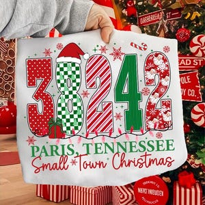 May include: A white Christmas-themed pillow with the numbers "38242" in red, green, and white patterns. The text "Paris, Tennessee Small Town Christmas" is below the numbers. Festive snowflakes and a Santa hat adorn the design.