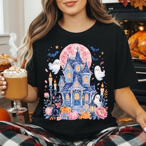 Puede incluir: Una camiseta negra con un estampado gr&aacute;fico colorido de una casa encantada azul, un gato negro, tres fantasmas blancos, calabazas, flores y velas sobre una gran luna rosa con murci&eacute;lagos y estrellas en el cielo nocturno.