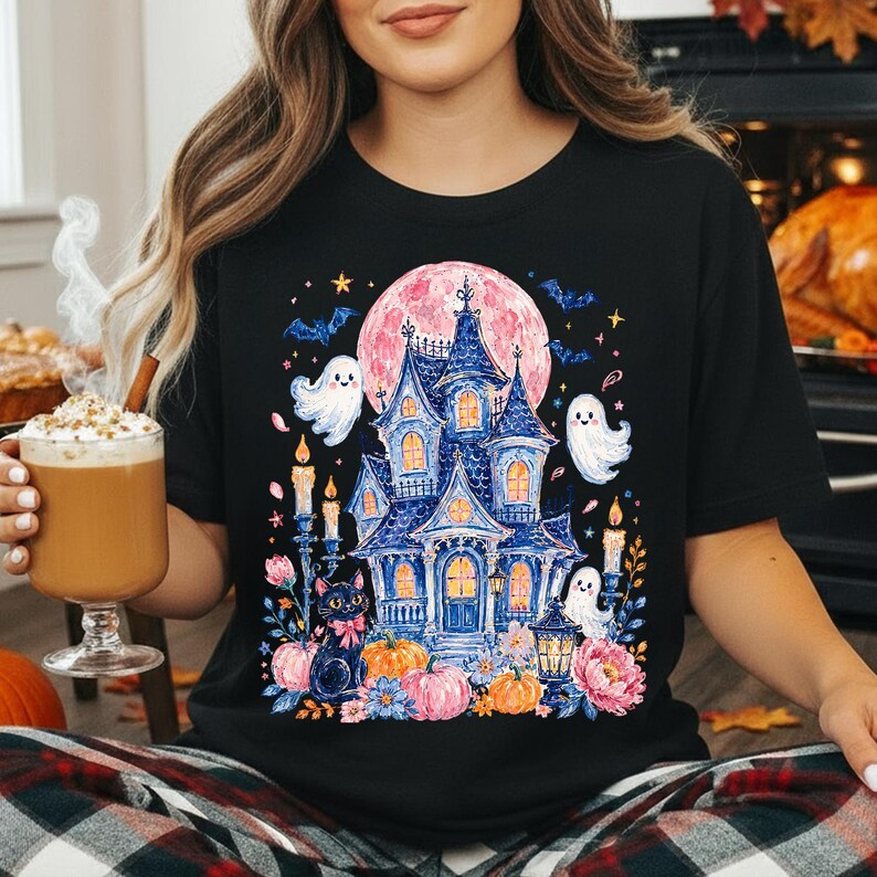 Puede incluir: Una camiseta negra con un estampado gr&aacute;fico colorido de una casa encantada azul, un gato negro, tres fantasmas blancos, calabazas, flores y velas sobre una gran luna rosa con murci&eacute;lagos y estrellas en el cielo nocturno.