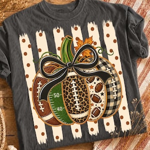 May include: A charcoal gray t-shirt featuring a large pumpkin graphic composed of various football-themed patterns, including leopard print, green field markings with 40 and 50 yard lines, and black and white plaid, all tied with a black bow.