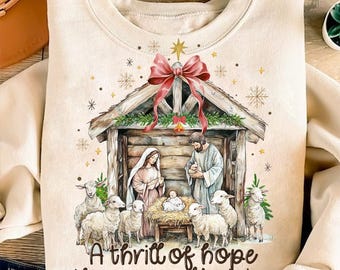 May include: A beige sweatshirt with a Christmas nativity scene illustration. The scene features a wooden manger with Mary, Joseph, and baby Jesus. There are sheep surrounding the manger and a red bow with a star above. The text "A thrill of hope the weary world rejoices" is printed below the scene.