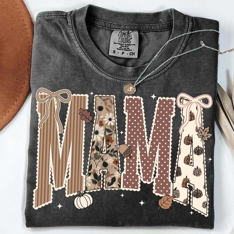 Peut inclure: Un t-shirt gris anthracite avec le mot MAMA en grandes lettres majuscules. Chaque lettre pr&eacute;sente un motif unique, notamment des rayures brunes, des fleurs, des pois bruns et des citrouilles &agrave; imprim&eacute; l&eacute;opard, avec des n&oelig;uds et des feuilles.