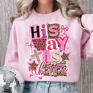 May include: A light pink sweatshirt with the phrase "His Way is Better" in a mix of fonts and patterns, including leopard print and gingham. The design also features a cross, stars, and hearts.