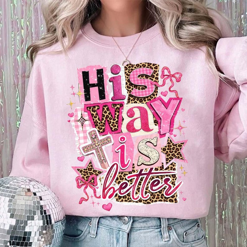 May include: A light pink sweatshirt with the phrase "His Way is Better" in a mix of fonts and patterns, including leopard print and gingham. The design also features a cross, stars, and hearts.