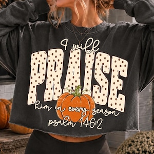 May include: A charcoal gray long-sleeve cropped sweatshirt featuring the text "I will PRAISE him in every season psalm 146:2" in white and beige polka dot lettering, with a central graphic of an orange pumpkin with a green stem.