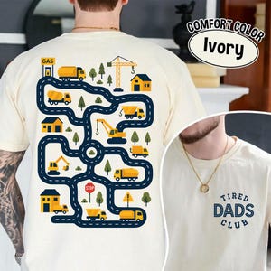 May include: Ivory-colored t-shirt with a construction-themed graphic on the back. The design features a road map with yellow trucks, houses, and a crane. The text "Tired Dads Club" is on the front.