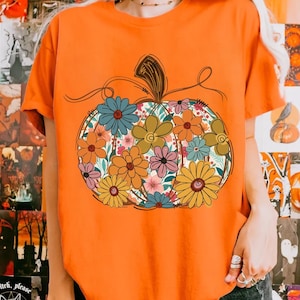 May include: An orange t-shirt with a floral pumpkin design. The pumpkin is filled with colourful flowers in shades of blue, orange, pink, and yellow. The design has a whimsical, hand-drawn style, ideal for autumn.