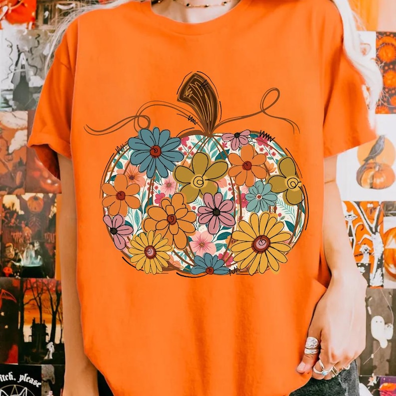 May include: An orange t-shirt with a floral pumpkin design. The pumpkin is filled with colourful flowers in shades of blue, orange, pink, and yellow. The design has a whimsical, hand-drawn style, ideal for autumn.