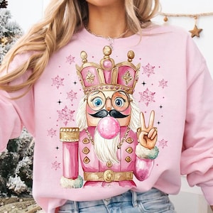 May include: Pink sweatshirt with a graphic of a pink and gold nutcracker wearing a crown and blowing a bubble. The nutcracker is holding up two fingers in a peace sign.