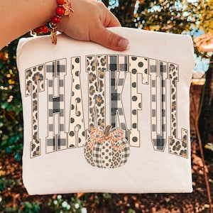 May include: A white t-shirt featuring the word THANKFUL in large block letters with a mix of leopard print, black and white buffalo plaid, and polka dot patterns. Below the text is a pumpkin graphic with matching patterns and a plaid bow.