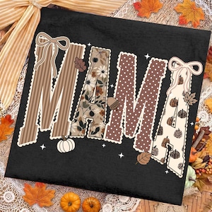 Peut inclure: Un t-shirt noir avec le mot MAMA en grandes lettres majuscules. Chaque lettre pr&eacute;sente un motif unique, notamment des rayures brunes, des fleurs, des pois bruns et des citrouilles &agrave; imprim&eacute; l&eacute;opard. Le design inclut des n&oelig;uds blancs et des &eacute;toiles.