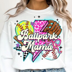 Puede incluir: Sudadera blanca con un dise&ntilde;o gr&aacute;fico colorido que presenta un b&eacute;isbol, una pelota de softbol, un bate y un guante. El texto "Ballpark Mama" se muestra en una fuente l&uacute;dica, con acentos rosas y negros.