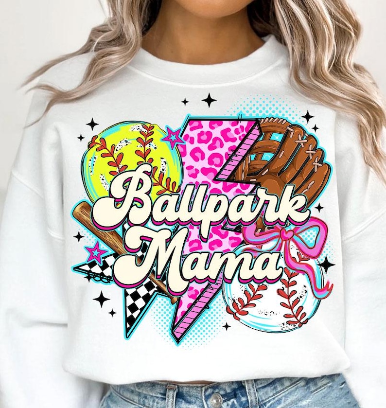 Puede incluir: Sudadera blanca con un dise&ntilde;o gr&aacute;fico colorido que presenta un b&eacute;isbol, una pelota de softbol, un bate y un guante. El texto "Ballpark Mama" se muestra en una fuente l&uacute;dica, con acentos rosas y negros.