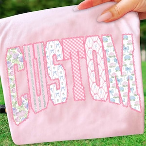 May include: A light pink fabric with the word "CUSTOM" in large, decorative letters. The letters have various patterns, including floral, gingham, and silver glitter stripes, all outlined with a scalloped pink trim.
