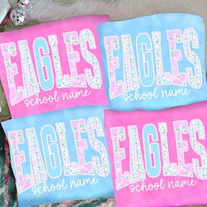 May include: Four t-shirts, two pink and two blue, with the word "EAGLES" in large, decorative lettering. The letters are outlined in white and feature floral and polka dot patterns. Below the word "EAGLES" is the text "school name" in a script font.