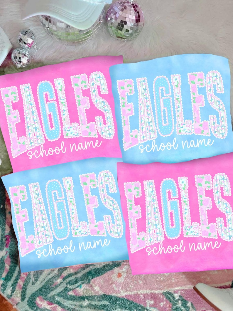 May include: Four t-shirts, two pink and two blue, with the word "EAGLES" in large, decorative lettering. The letters are outlined in white and feature floral and polka dot patterns. Below the word "EAGLES" is the text "school name" in a script font.
