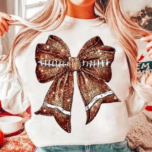 Könnte beinhalten: Weißer Pullover mit einer braunen Glitzer Schleife mit weißen Nähten auf der Schleife. Die Schleife ist in Form eines Fußballs.