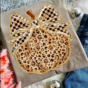 May include: A tan-colored t-shirt featuring a pumpkin design with a leopard print pattern and skull accents. The pumpkin has a large bow with a black and white checkered pattern. The design is outlined with gold glitter.